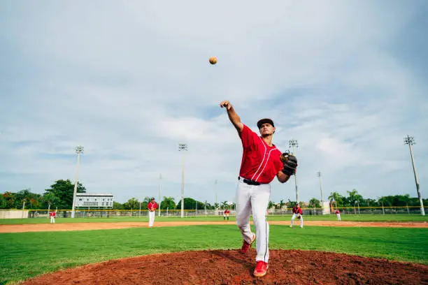 Low angle view of young Hispanic baseball pitcher delivering a pitch from the mound with fielders in background.