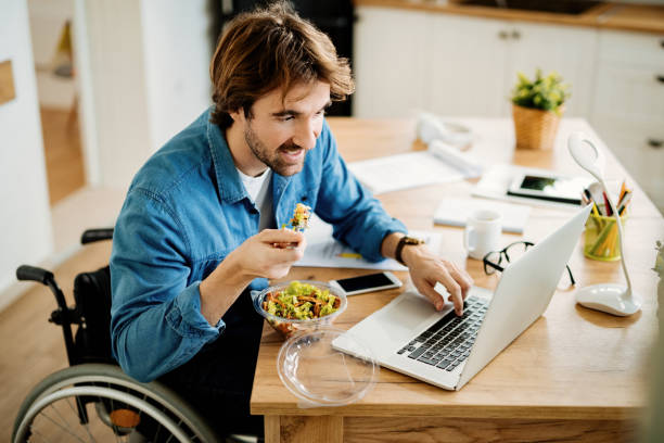 lavoratore freelance sorridente in sedia a rotelle che lavora su laptop mentre mangia a casa. - man eating foto e immagini stock