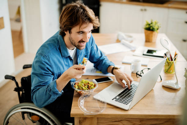 uśmiechnięty niezależny pracownik na wózku inwalidzkim pracujący na laptopie podczas jedzenia w domu. - healthy eating healthcare and medicine healthy lifestyle people zdjęcia i obrazy z banku zdjęć