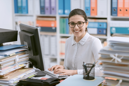 Young smiling secretary working at office desk and stacks of paperwork