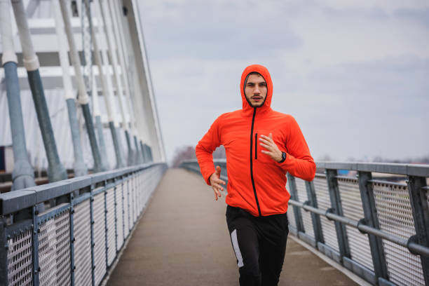 istock Handsome young athlete running fast along big modern bridge in orange windbreaker jacket 1212160282