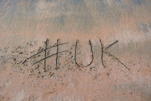 Image of hashtag tag writing on sunny beach in the soft sand, light by the early morning sunshine. Written using a stick by hand, with UK words as capital letters by the gentle lapping waves of the sea on the seashore. Picture taken at Palolem Beach, Goa, India, during the early morning golden hour without tourists on holiday vacation / beachgoers. Concept photo of UK handwriting in golden sand with modern hashtag prefix imagen de hashtag tag etiqueta escritura en playa soleada con palabra del reino unido escrita en arena por mar, palolem beach, goa, india, concepto de la foto de las redes sociales de escritura a mano en arena dorada con el hashtag moderno prefiix