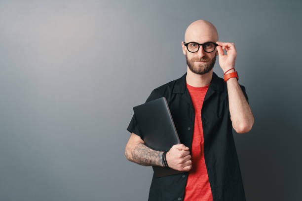 A Young bearded man with glasses holding laptop under arm in the studio on gray background stock photo