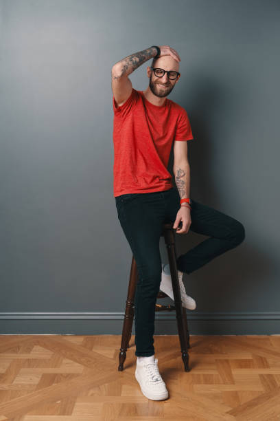 istock Young smiling man with beard and glasses sitting on a tall chair on gray studio background 1212161646