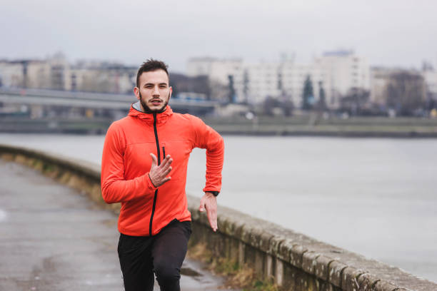 istock Handsome young athlete man running fast along river in orange windbreaker jacket 1212163111