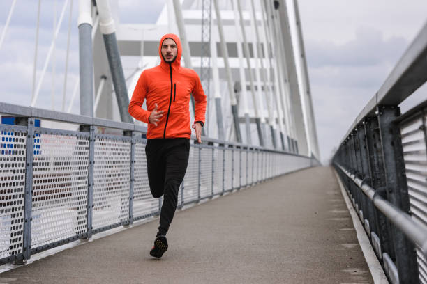 istock Handsome young athlete running fast along big modern bridge in orange windbreaker jacket 1212164546