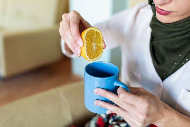 An Unhealthy Muslim woman with cold or flu is sitting under the blanket on the sofa in the living room drinking cup of tea with lemon. A woman of Middle east ethnicity with seasonal infections, flu, allergy taking a hot drink at home.