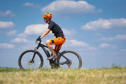 45 year old man on his e-bike mountainbike cycling down a dirt road in his bikewear black bike orange bikedress and helmet in front of blue sky