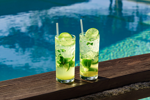 Two fresh and cold mojitos with ice cubes, lime slices and mint leaves on a turquoise pool background