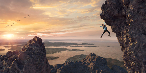 A wide angle panoramic image of a female free climber ascending a steep rock face. The climber is wearing black leggings and climbing top and hangs by one hand from the rock. She is at high altitude above rocky peaks in a coastal location at sunset.