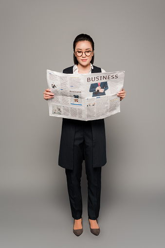 young businesswoman in eyeglasses reading newspaper on grey background