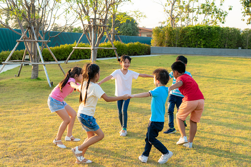 Large group of happy Asian smiling kindergarten kids friends holding hands playing and dancing play roundelay and stand in circle in the park on the green grass on sunny summer day.