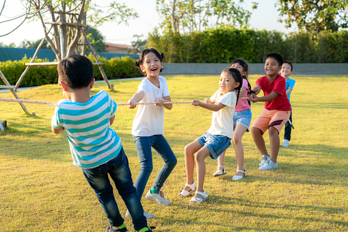 Group of happy young Asian children playing tug of war or pull rope together outside in city park playground in summer day. Children and recreation concept.
