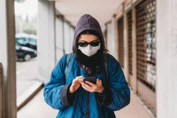 Portrait of a young hip woman in the city with flu mask using a mobile phone. She's protecting herself from viruses. Closed shops in the background.