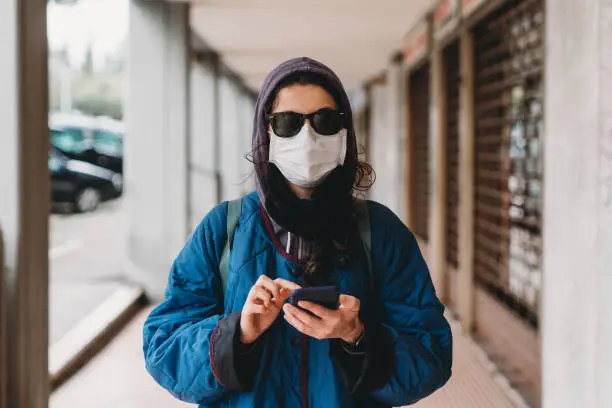 Portrait of a young hip woman in the city with flu mask using a mobile phone. She's protecting herself from viruses. Closed shops in the background.