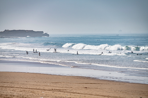 Atlantic Ocean at Taghazout, a tourist area in Morocco that is receiving considerable investment in new hotels, apartments etc. This shows the beach with surf up and many tourists swimming and surfing in the background.