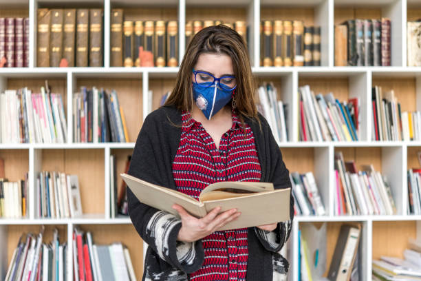 コロナ ウイルス。コロナウイルスのマスクを持つ図書館の学生。ビジネスウーマンは、自分の健康を保護し、世話をするためにマスクを着用します。 - bookshelf beautiful intelligence learning ストックフォトと画像