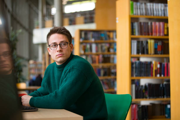 Young Adult Man Sitting By a Table in Library and Looking Around stock photo