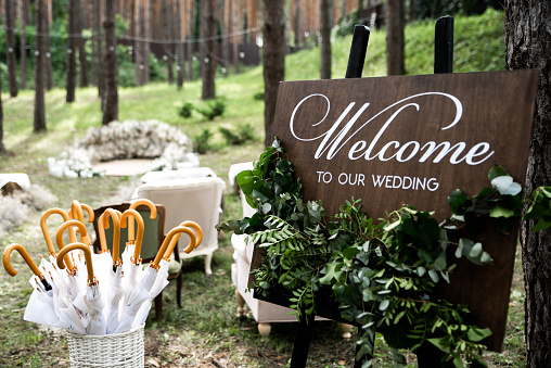 Rustic style wedding decor. Outdoor wooden welcome board decorated with green branches, basket with umbrellas in case of rain
