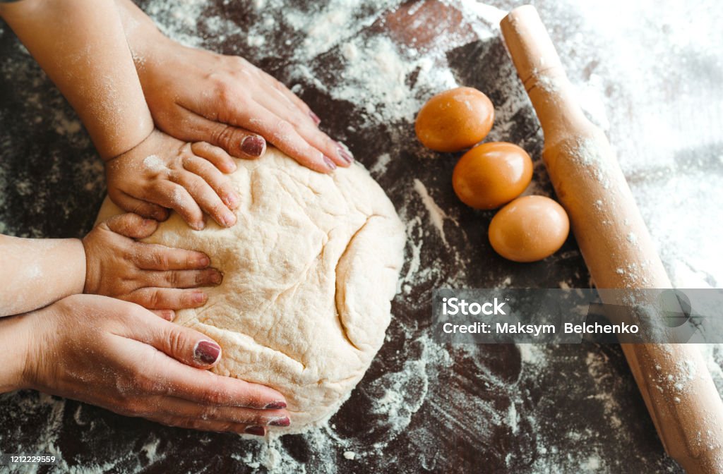 Hands of mother and son kneading a dough together. Photo. Hands of mother and son kneading a dough together. Mother and child hands prepares the dough with flour on dark wooden table. Bakery background. Cooking concept. Dough Stock Photo
