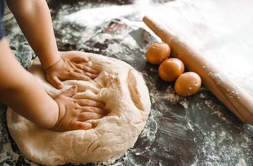 Photo. Little child preparing dough for baking. Small hands kneading dough and rolled dough with a rolling pin on the table. Kids hands, some flour, wheat dough and rolling-pin on the dark table. Cooking.