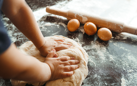 photo. Little child preparing dough for baking. Small hands kneading dough and rolled dough with a rolling pin on the table. Kids hands, some flour, wheat dough and rolling-pin on the dark table. Cooking.