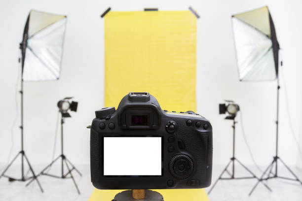 istock Camera in a photo studio with yellow backdrop and light equipment. White screen mockup 1212239795