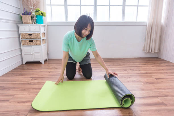 リビングルームで自宅でワークアウトした後、緑のヨガやフィットネスマットを折りたたむ魅力的なアジアの女の子のクローズアップ。健康的な生活は、フィット感の概念を保ちます。水平� - yoga business women living room ストックフォトと画像