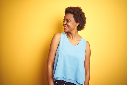 Beautiful african american woman wearing elegant shirt over isolated yellow background looking away to side with smile on face, natural expression. Laughing confident.