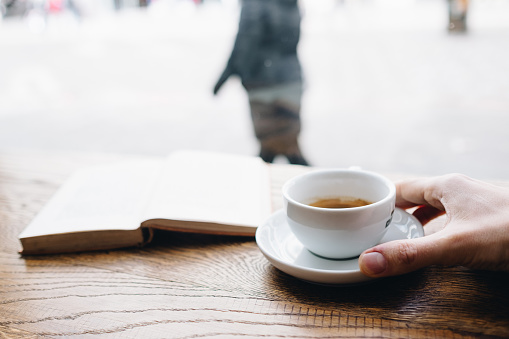 Human hand holding black coffee on saucer with vintage book on background on the wooden table next to big coffee shop window and looking to passerby street walkers