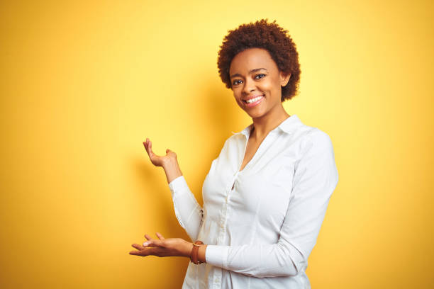 donna d'affari afroamericana su sfondo giallo isolato invitante ad entrare sorridente naturale a mano aperta - tentazione foto e immagini stock