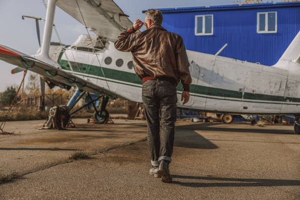 Back view of fashionable pilot in leather jacket and sunglasses near plane outdoors stock photo