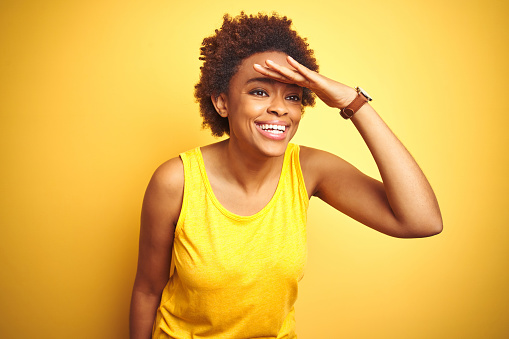 Beauitul african american woman wearing summer t-shirt over isolated yellow background very happy and smiling looking far away with hand over head. Searching concept.