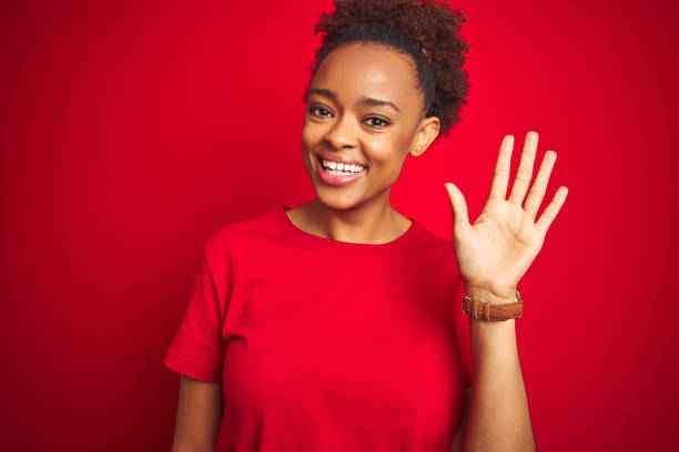 Young beautiful african american woman with afro hair over isolated red background Waiving saying hello happy and smiling, friendly welcome gesture Young beautiful african american woman with afro hair over isolated red background Waiving saying hello happy and smiling, friendly welcome gesture hello single word photos stock pictures, royalty-free photos & images