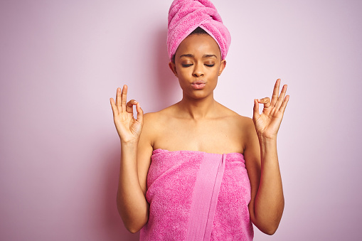 African american woman wearing shower towel after bath over pink isolated background relax and smiling with eyes closed doing meditation gesture with fingers. Yoga concept.