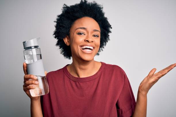リフレッシュメントのための巻き毛のボトルの水を飲む若いアフリカ系アメリカ人のアフロの女性は、満面の笑みと手を挙げて叫んで勝利を祝う勝者の表情 - big bottle ストックフォトと画像