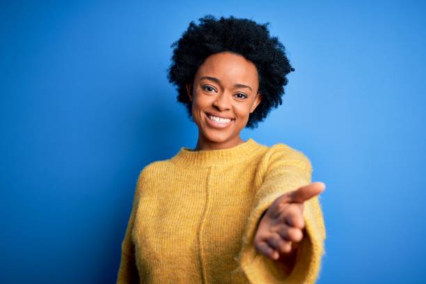 黄色のカジュアルセーターを着た巻き毛の若い美しいアフリカ系アメリカ人のアフロ女性は、助けと受け入れを与える手のひらの手を提供する陽気な笑顔をしています。 - african descent women american culture human hair ストックフォトと画像