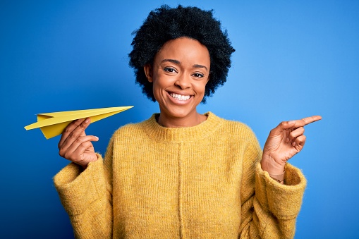 Young African American woman with curly hair holding paper airplane over blue background very happy pointing with hand and finger to the side