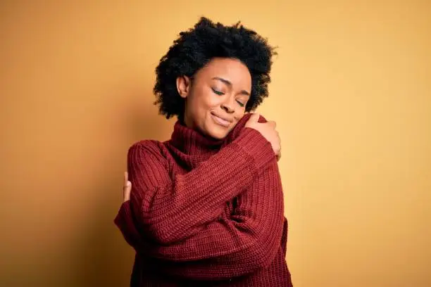 Young beautiful African American afro woman with curly hair wearing casual turtleneck sweater Hugging oneself happy and positive, smiling confident. Self love and self care