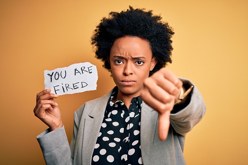 Young African American afro woman with curly hair holding paper with you are fired message with angry face, negative sign showing dislike with thumbs down, rejection concept