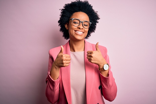 Joven hermosa afroamericana afro mujer de negocios con el pelo rizado usando chaqueta rosa signo de éxito haciendo gesto positivo con la mano, pulgares hacia arriba sonriendo y feliz. Expresión alegre y gesto ganador. photo