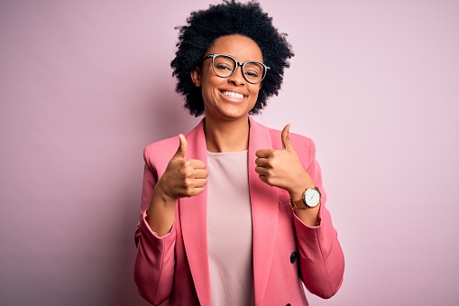 Young beautiful African American afro businesswoman with curly hair wearing pink jacket success sign doing positive gesture with hand, thumbs up smiling and happy. Cheerful expression and winner gesture.