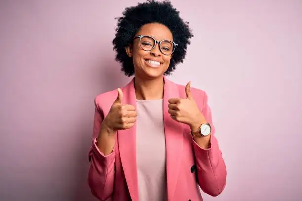 Photo of Young beautiful African American afro businesswoman with curly hair wearing pink jacket success sign doing positive gesture with hand, thumbs up smiling and happy. Cheerful expression and winner gesture.