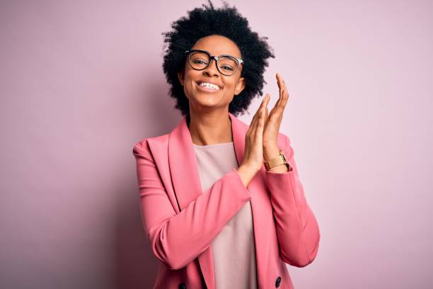 junge schöne afrikanische afroamerikanische afro geschäftsfrau mit lockigen haaren trägt rosa jacke klatscht und applaudiert glücklich und fröhlich, lächelnd stolze hände zusammen - applauding stock-fotos und bilder