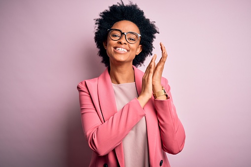Joven hermosa afroamericana afro mujer de negocios con el pelo rizado usando chaqueta rosa aplaudiendo y aplaudiendo felices y alegres, sonriendo manos orgullosas juntos photo