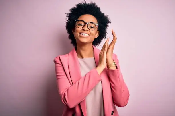 Young beautiful African American afro businesswoman with curly hair wearing pink jacket clapping and applauding happy and joyful, smiling proud hands together