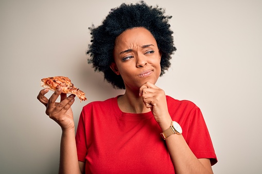 Young African American afro woman with curly hair eating slice of delicious Italian pizza serious face thinking about question, very confused idea