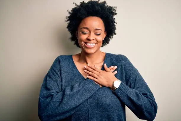 Photo of Young beautiful African American afro woman with curly hair wearing casual sweater smiling with hands on chest with closed eyes and grateful gesture on face. Health concept.