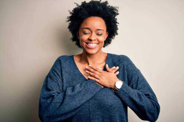 young beautiful african american afro woman with curly hair wearing casual sweater smiling with hands on chest with closed eyes and grateful gesture on face. health concept. - peito imagens e fotografias de stock