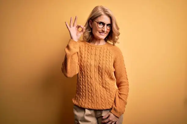 Middle age beautiful blonde woman wearing casual sweater and glasses over yellow background smiling positive doing ok sign with hand and fingers. Successful expression.